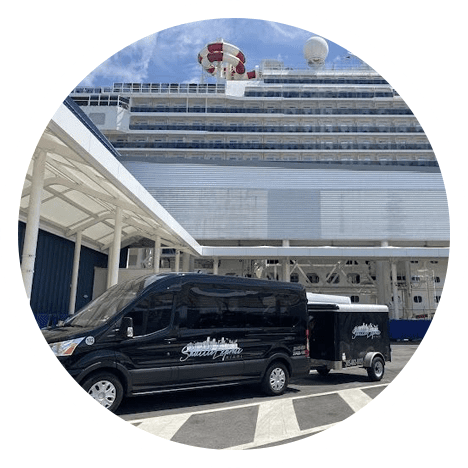 Black shuttle vans parked in front of a large cruise ship docked at a port, under a clear sky.